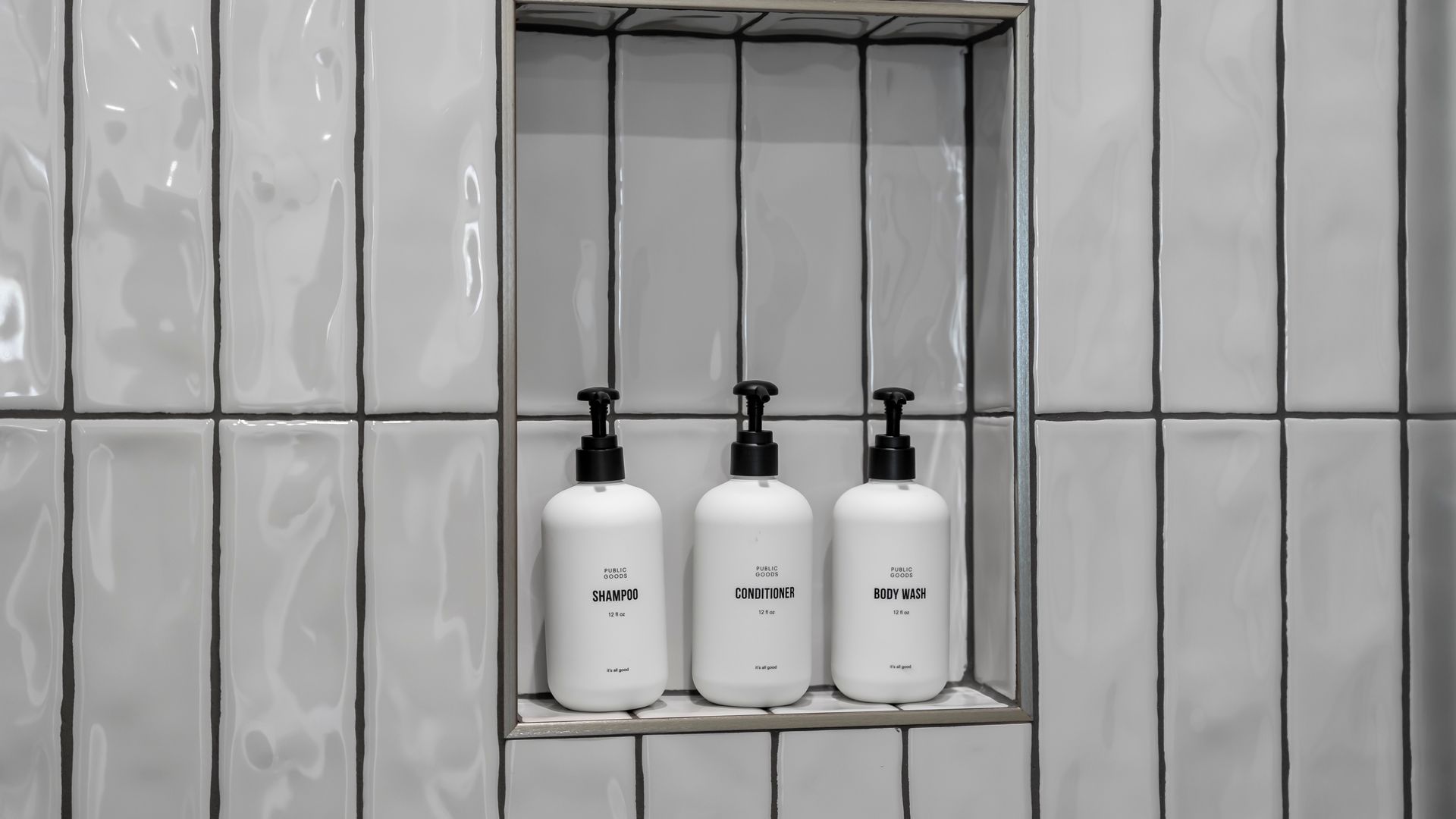 Three white soap bottles in a shower shelf, set against white vertical tiled wall.