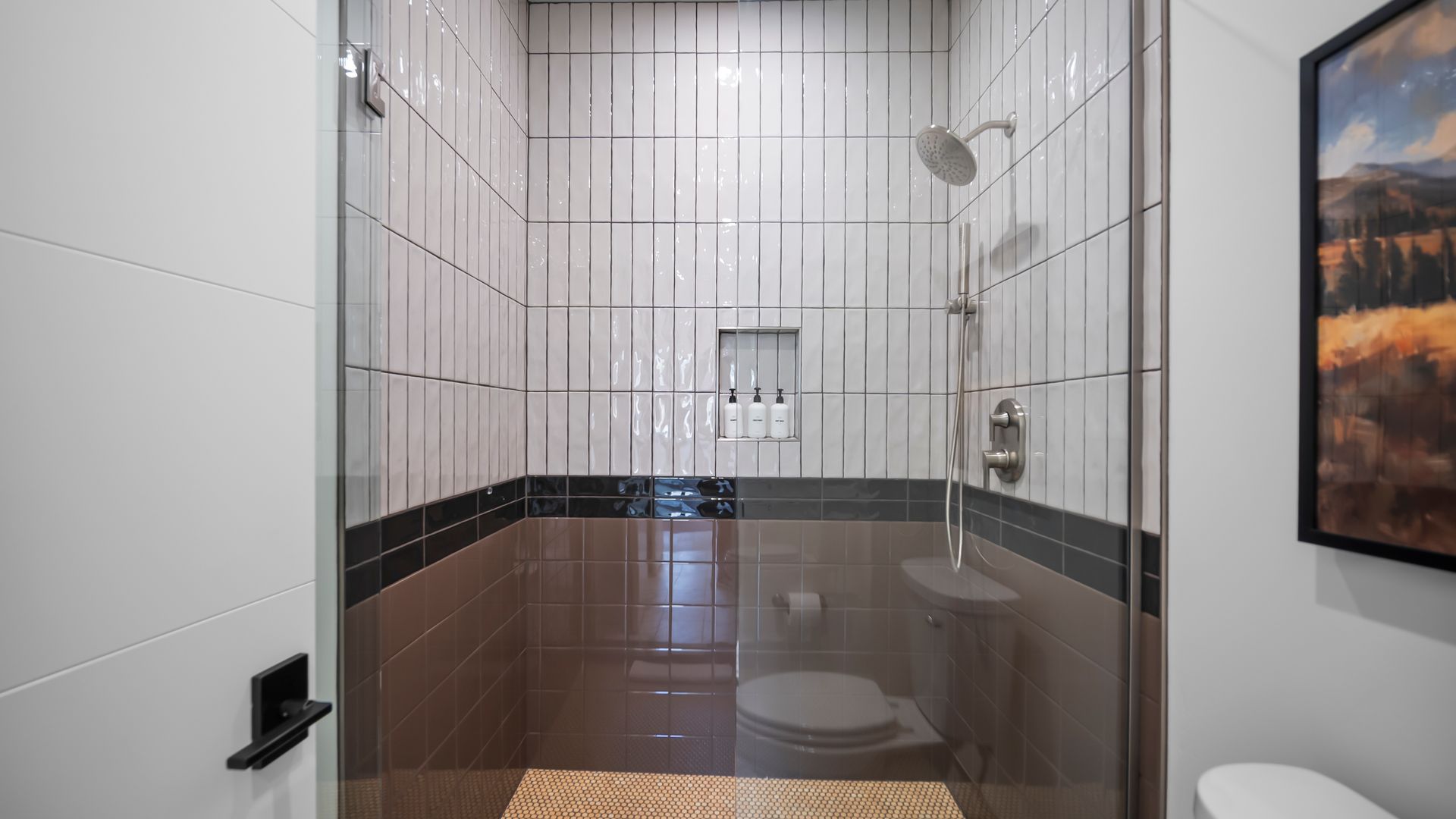 Bathroom shower with glass door, white and brown tiled walls, silver showerhead, and toilet.