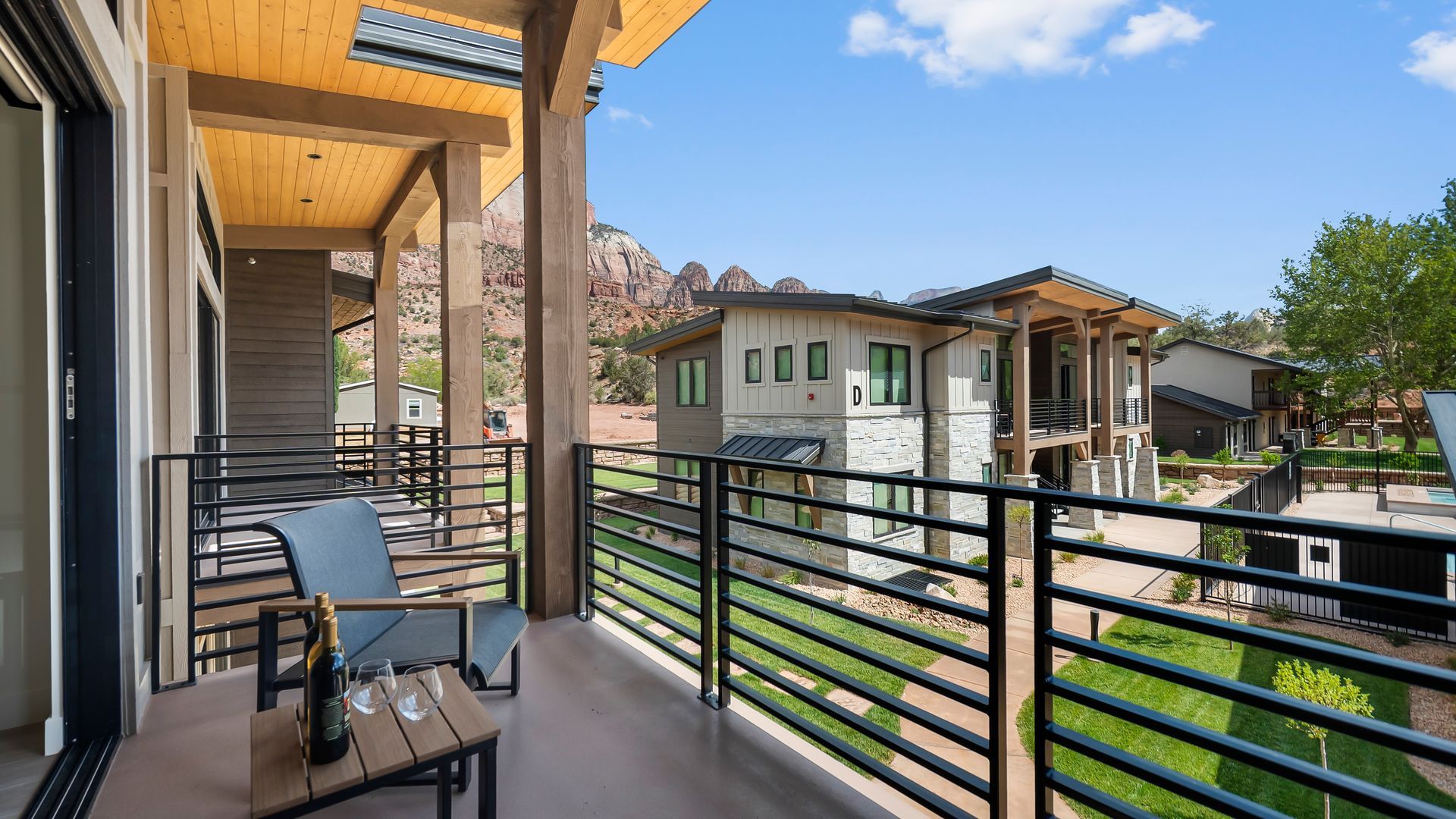 Balcony with chair and table, overlooking a green lawn and modern houses, against a mountainous backdrop.