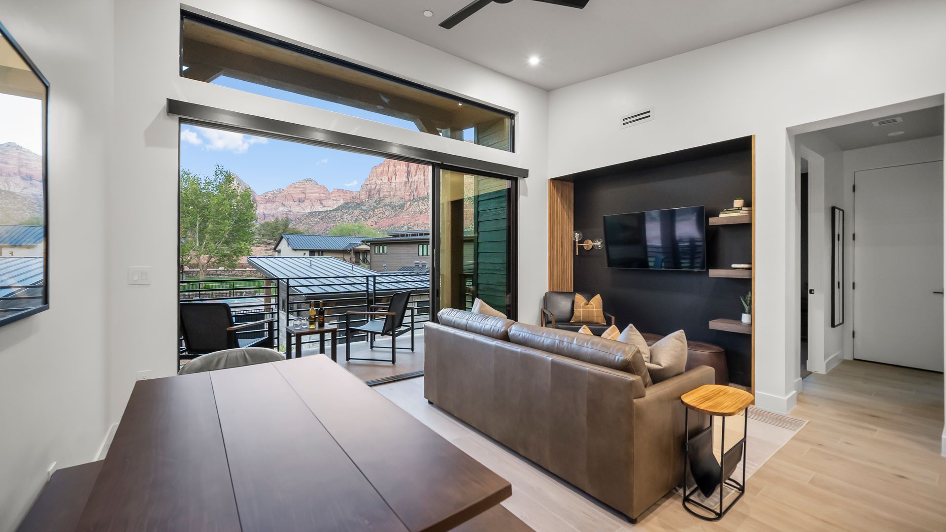 Modern living room with open door to patio, mountain view, brown leather sofa, and dark accent wall.