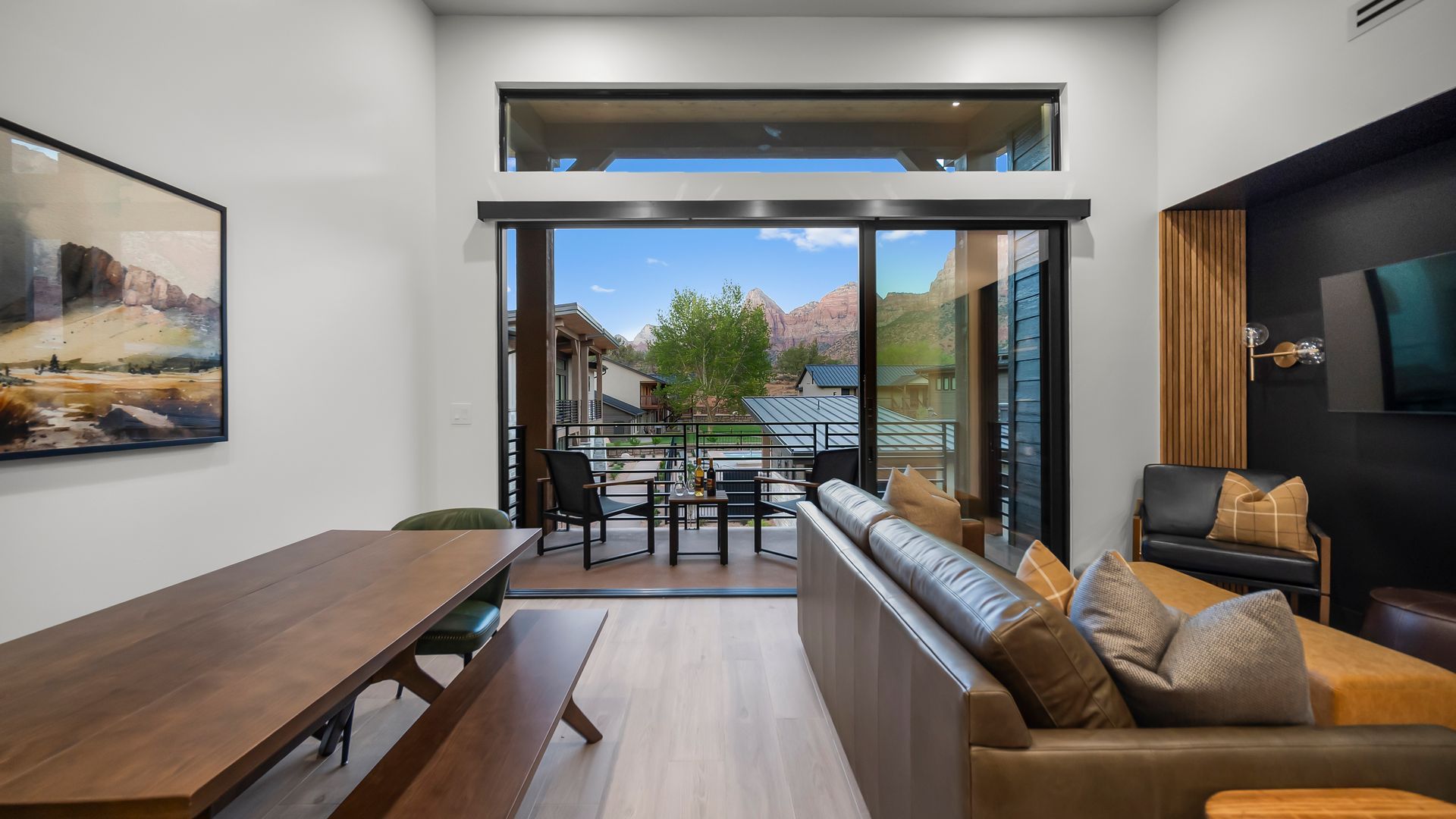 Living room with open doors to a patio overlooking greenery. Brown leather sofa, wooden table, and art piece.