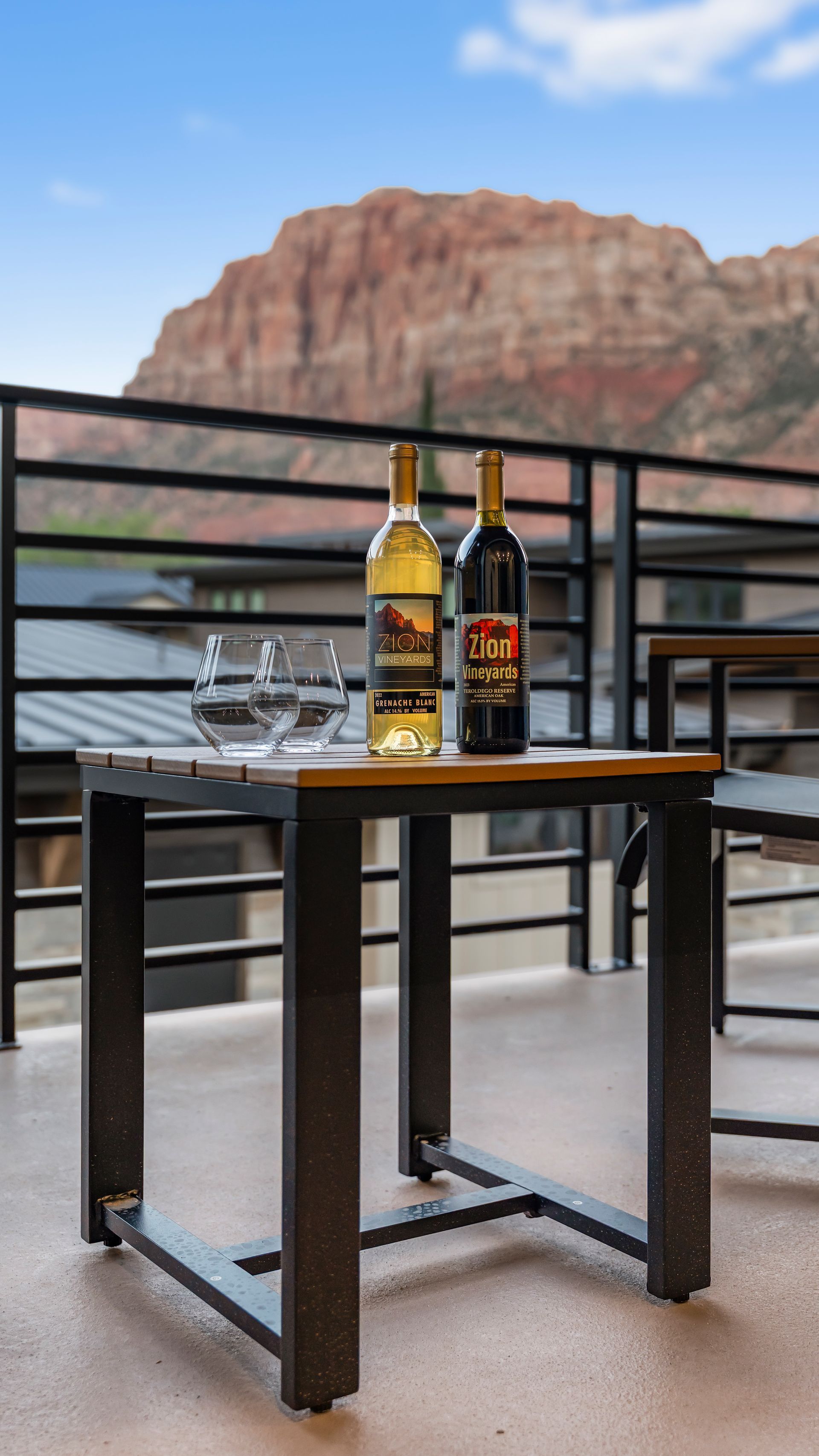 Two wine bottles on a table with a mountain backdrop. A glass of water sits nearby.