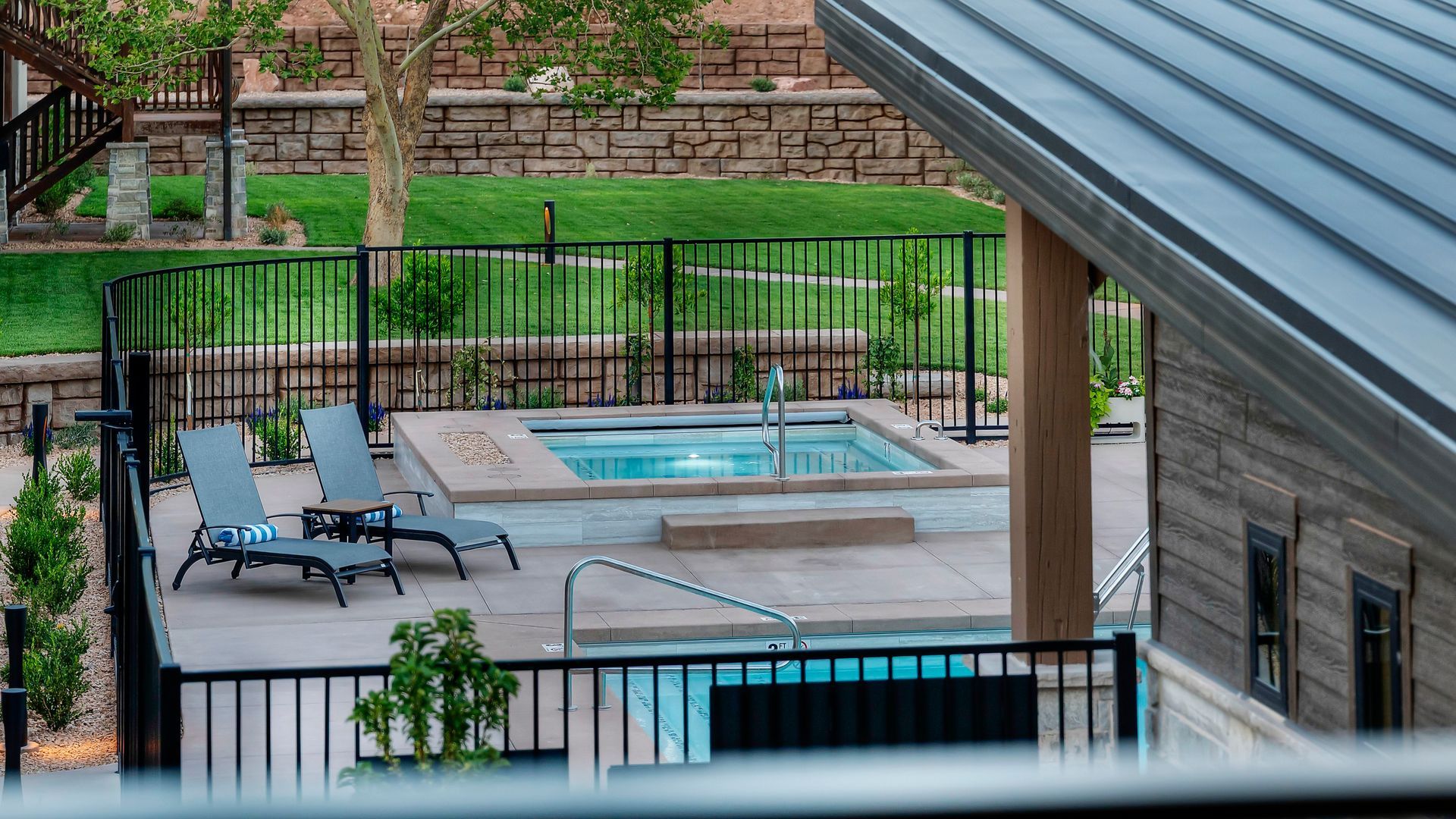 Hot tub and pool area, fenced with black metal. Lounging chairs, landscaping, and building facade visible.