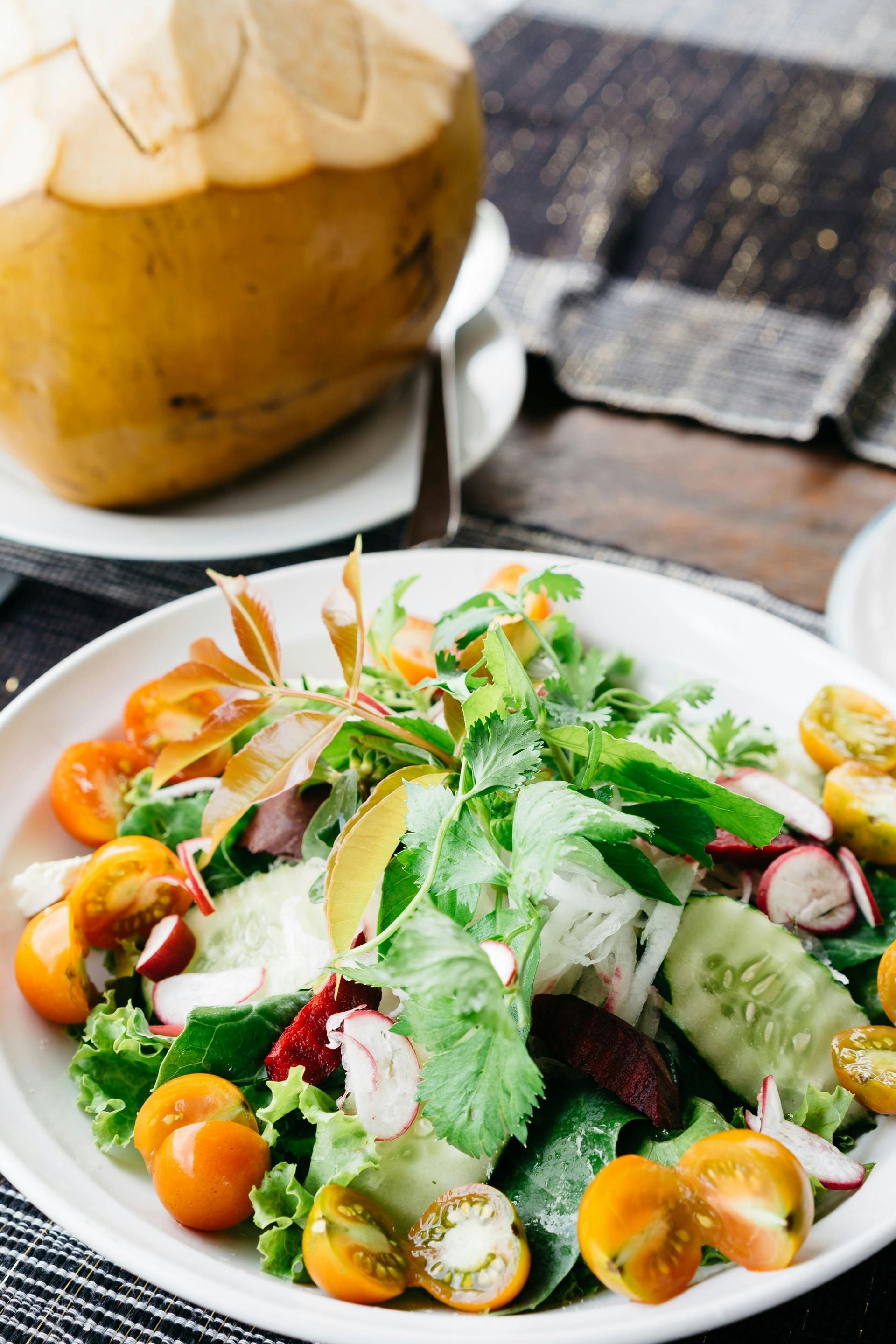 Fresh salad with tomatoes, radishes, and greens, beside a coconut on a wooden table.