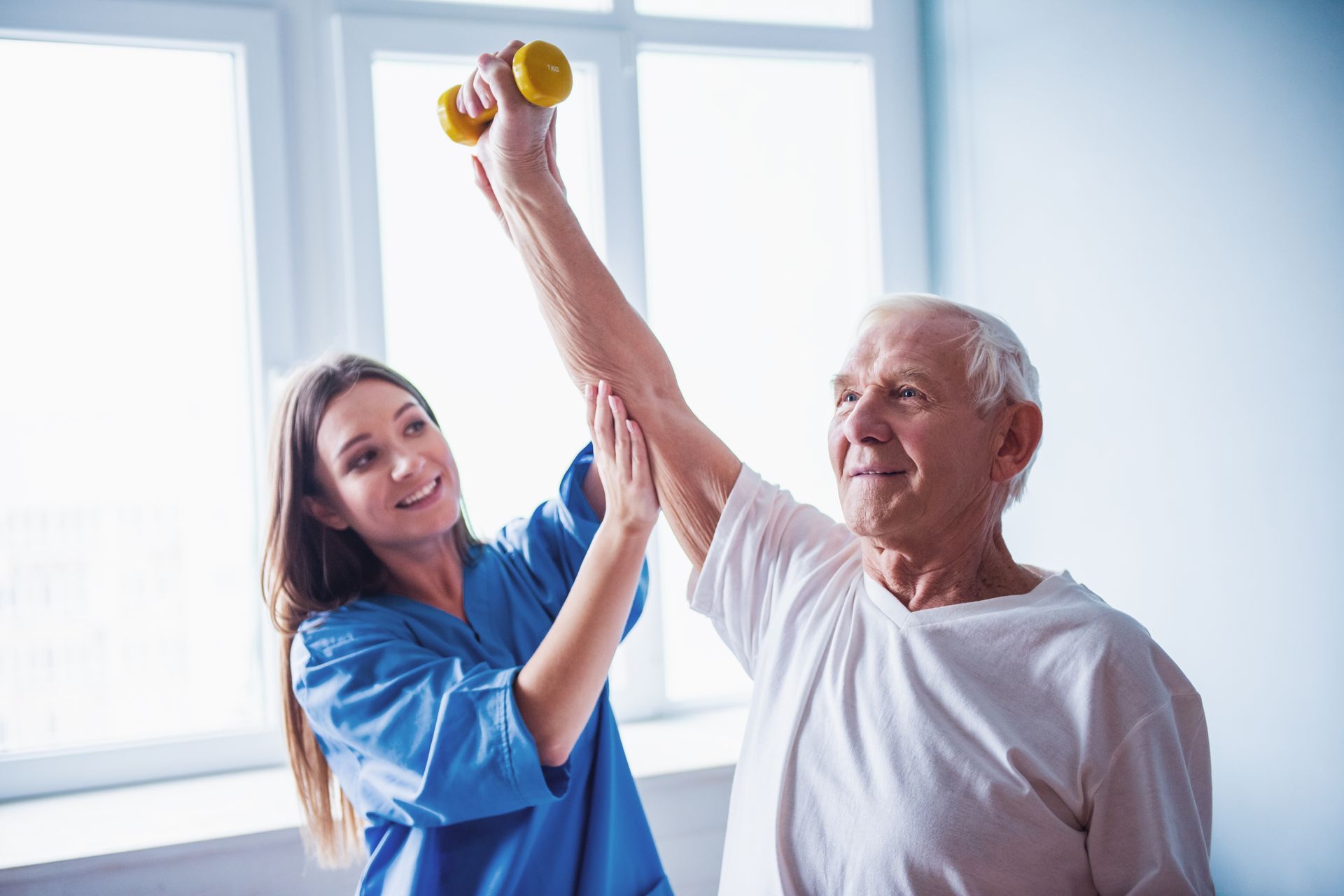 Woman in blue scrubs helps an elderly man raise a small dumbbell in physical therapy.