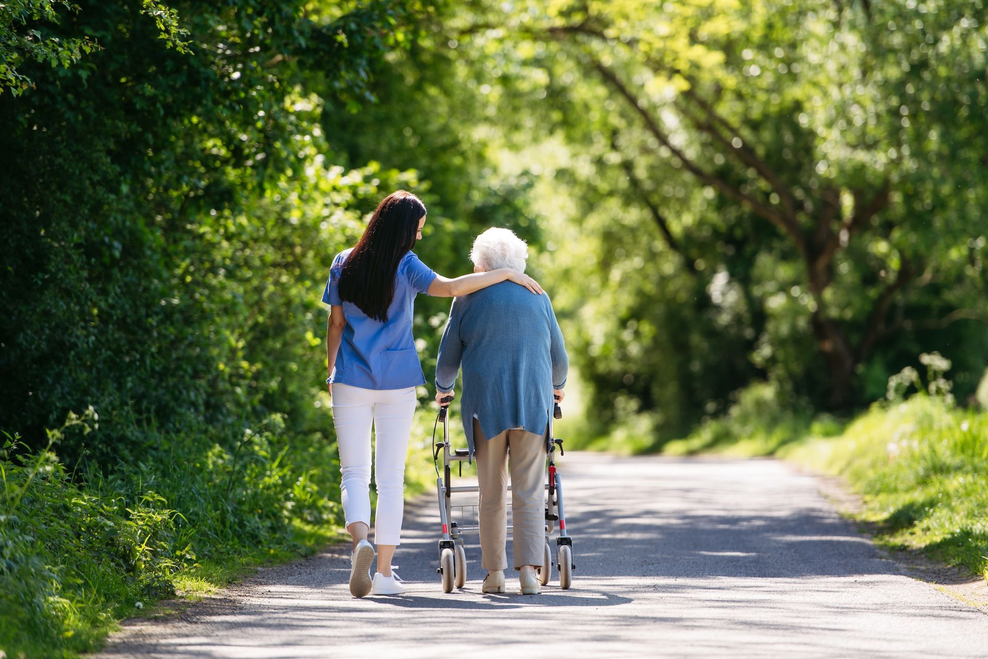 A caregiver assists an elderly woman using a walker along a tree-lined path.