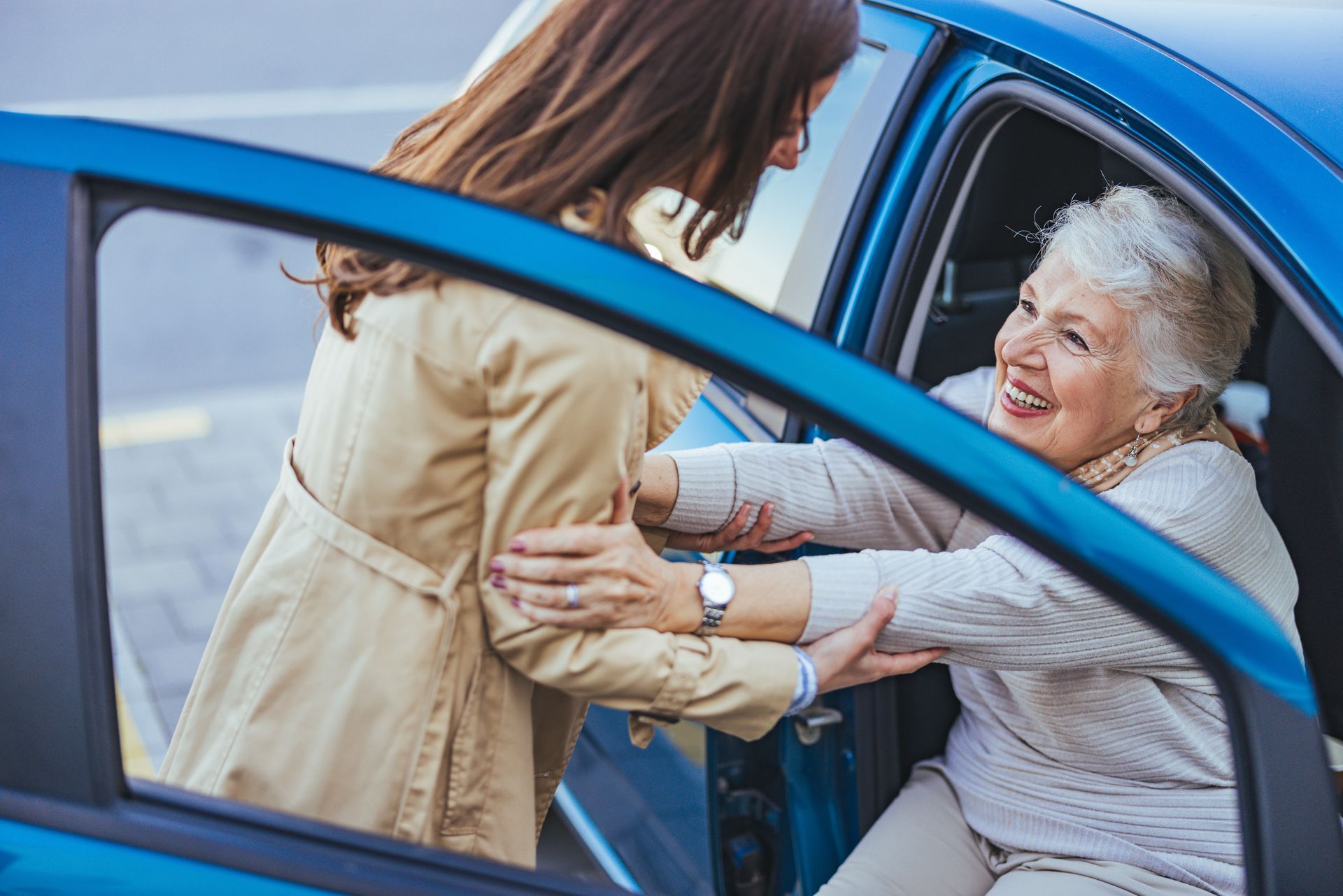 Woman helping elderly woman get out of a blue car; both smiling.