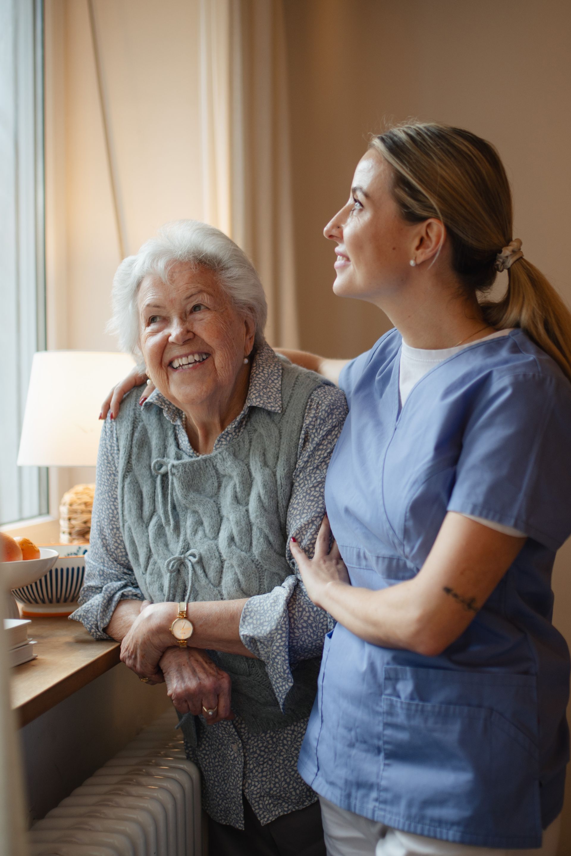 Elderly woman smiles, arm-in-arm with a caregiver. Both look out a window. Soft, natural light in a home setting.
