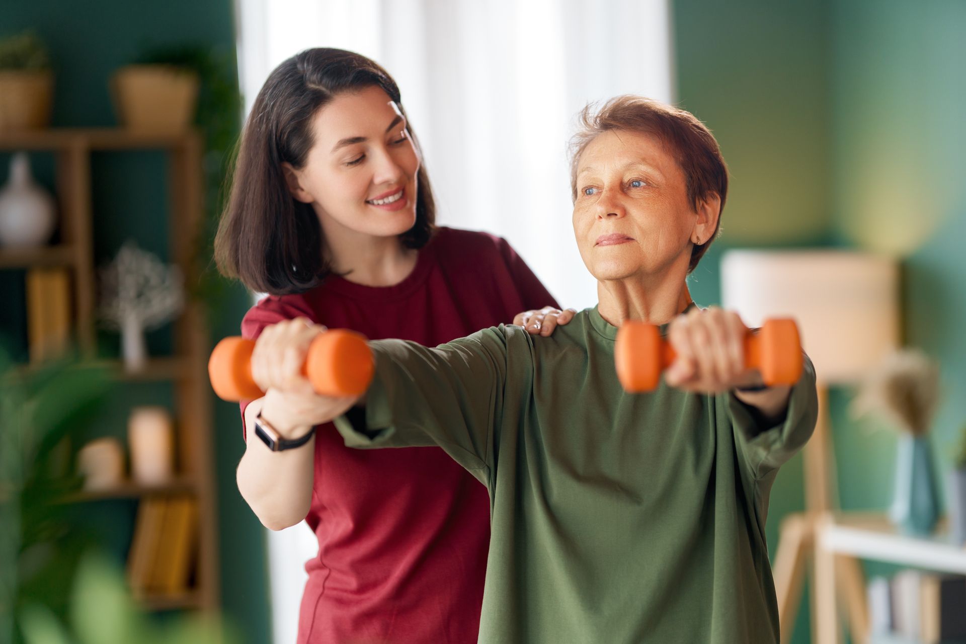 Woman assists elderly woman with dumbbell exercises. Indoors, green and red, smiling.