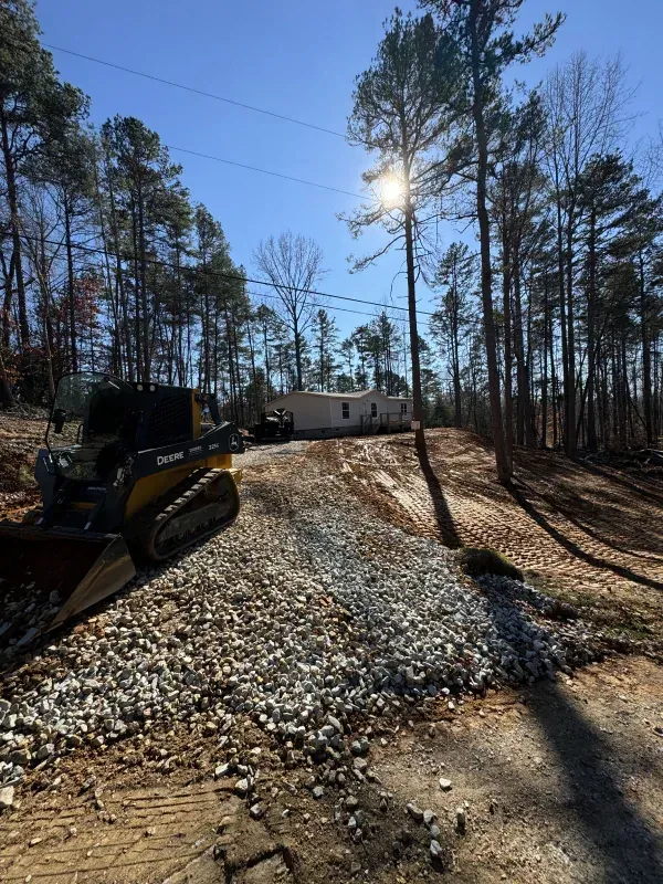 A gravel driveway being graded by a small construction vehicle leads to a house in a wooded area under a sunny sky.