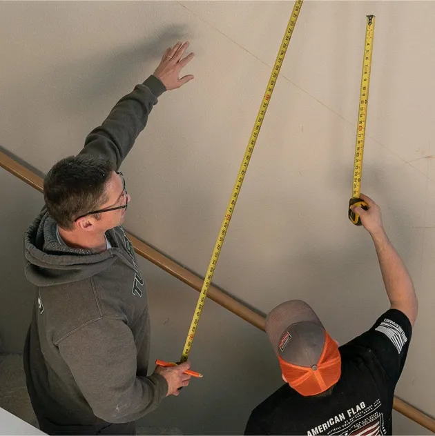 Two men are measuring a wall with tape measures
