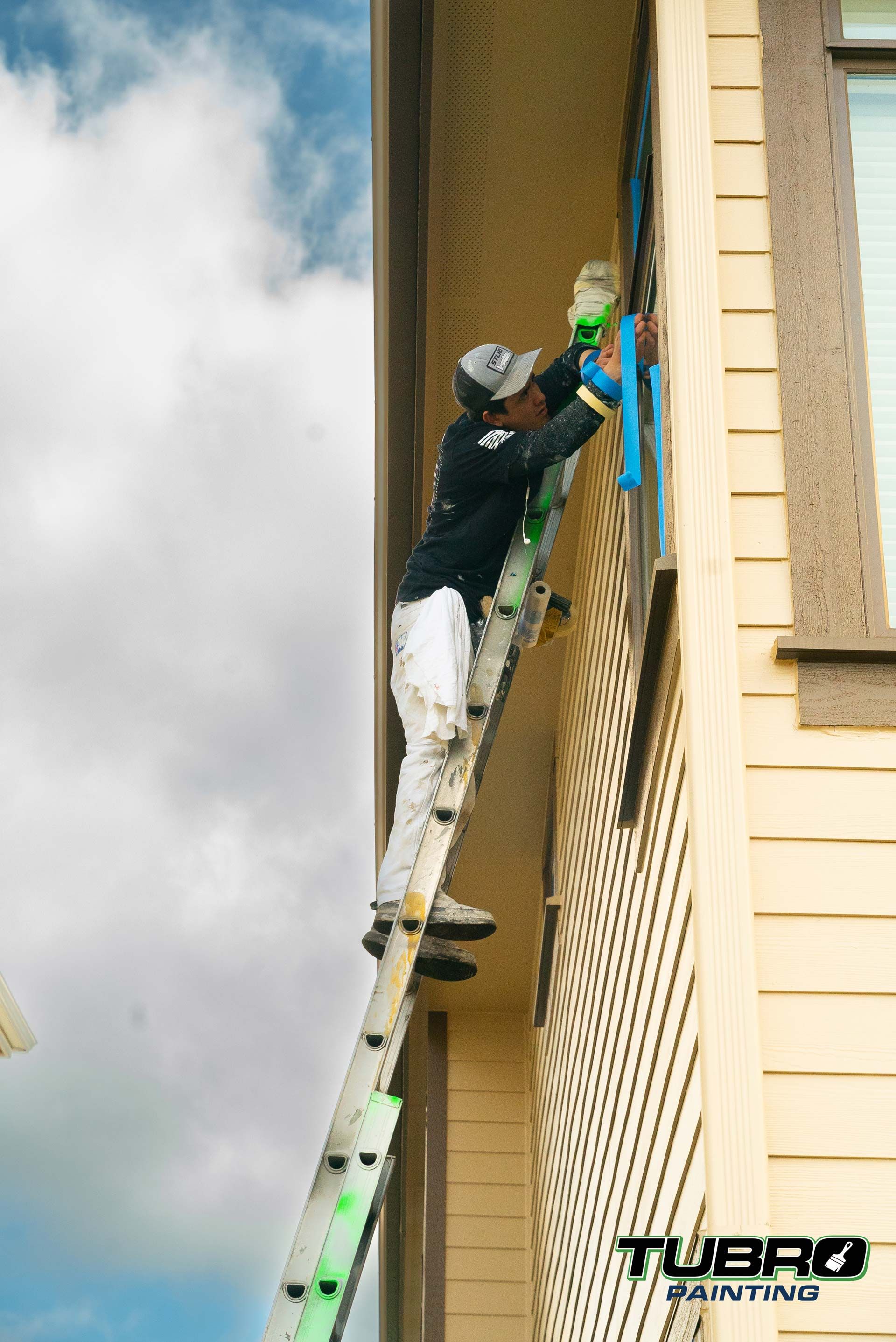 A man is standing on a ladder painting the side of a building.