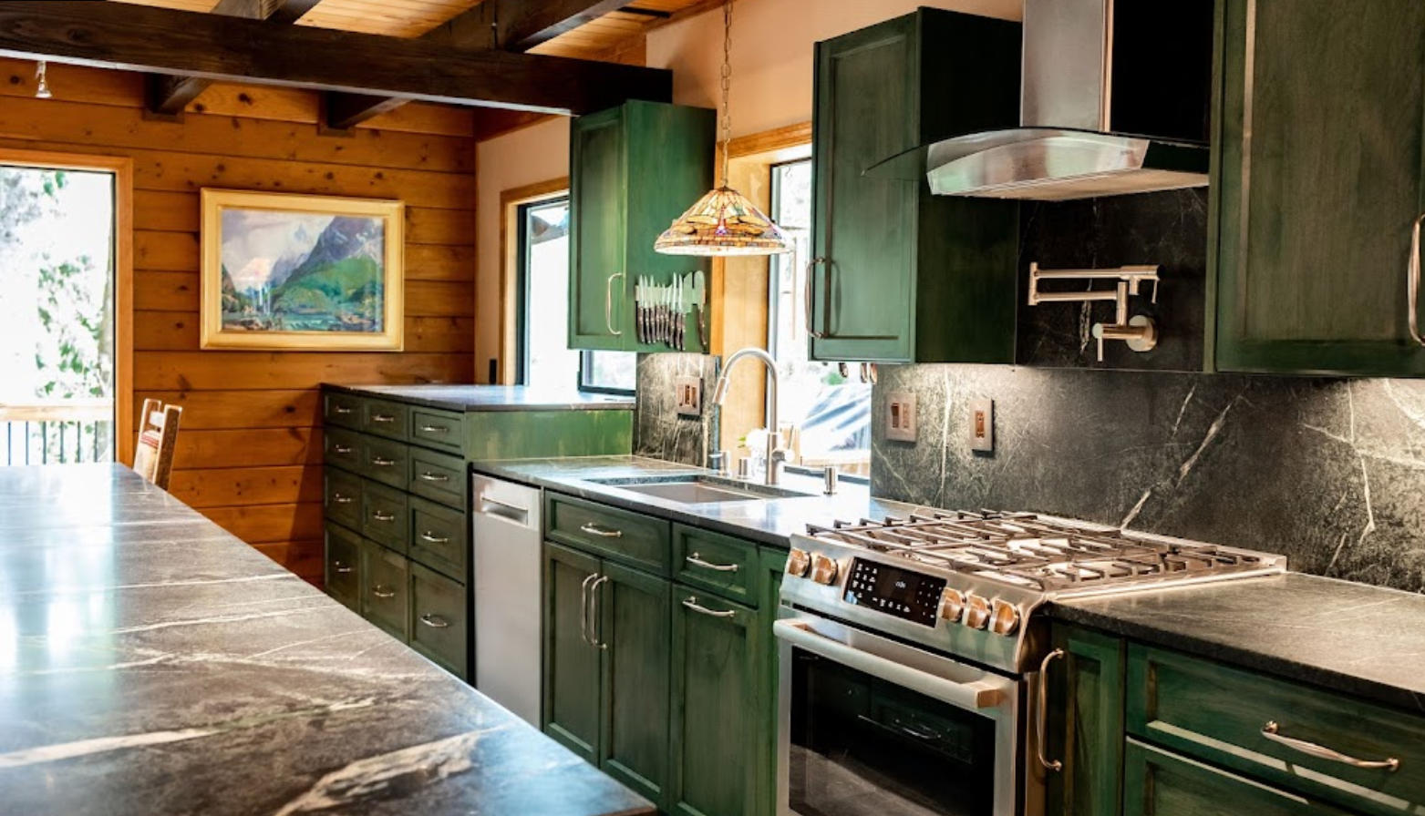 A kitchen with green cabinets , a stove , a sink , and a painting on the wall.