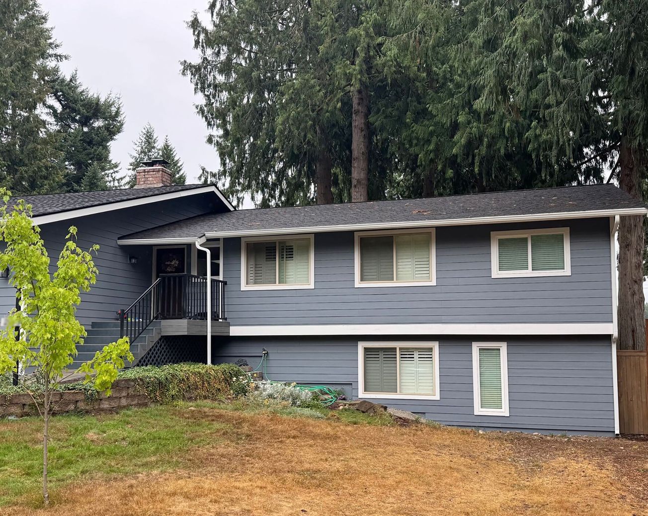 Gray kitchen with island, cabinets, and appliances. Light wood floor, stairs in background. Gray two-story house with white trim, windows, and a dark roof; set in front of trees, on a slightly sloped yard.