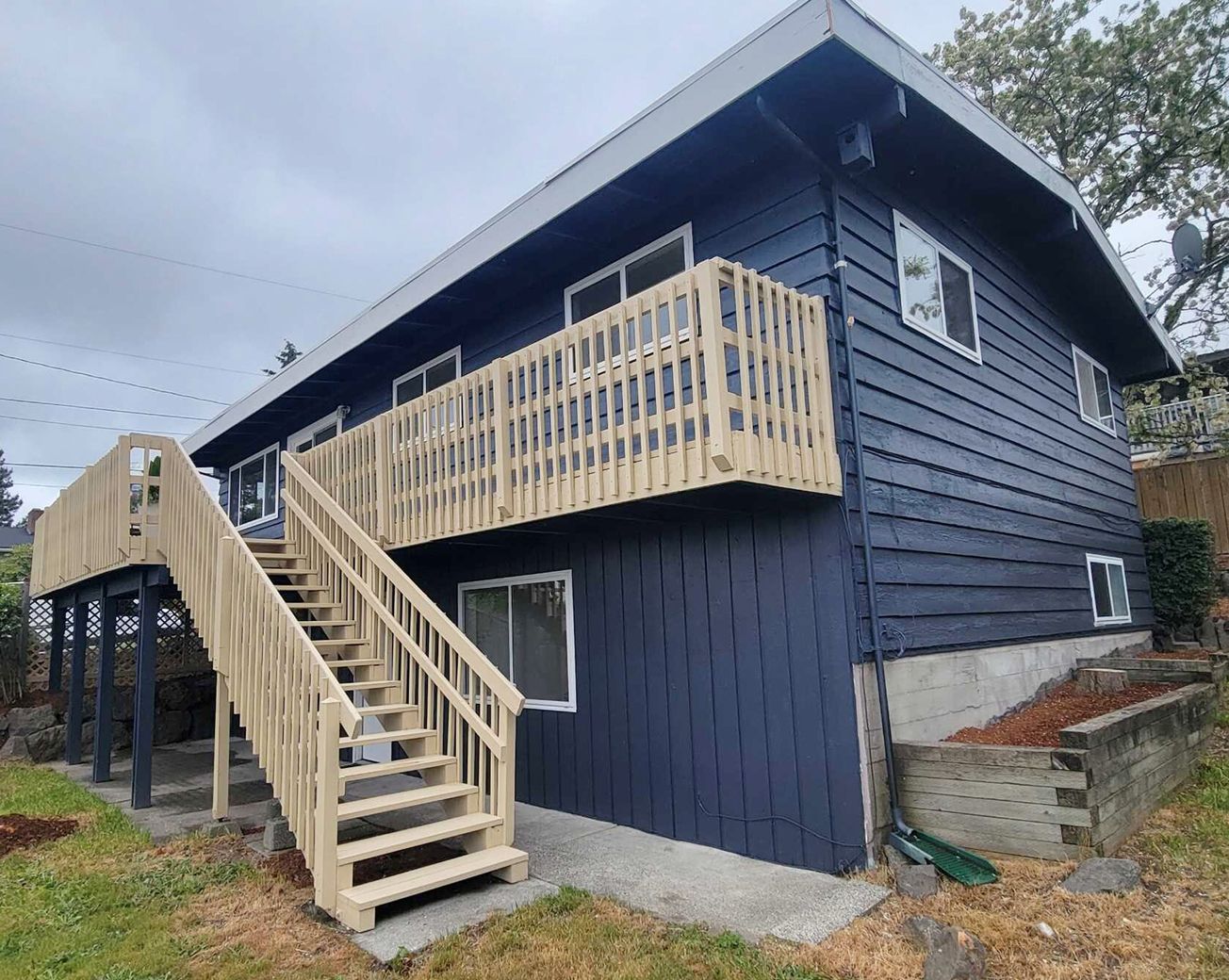 Two-story house with navy siding and wooden deck/stairs, overcast sky.
