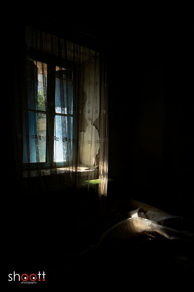 Licht stroomt door een raam met doorschijnende, versleten gordijnen een donkere, schaduwrijke kamer binnen en verlicht een gestructureerd oppervlak eronder.