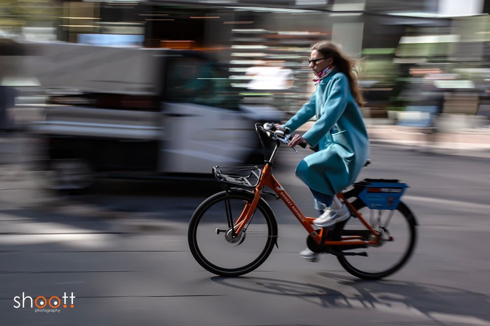 Een persoon in een turquoise jas rijdt op een oranje fiets door een stadsstraat, vastgelegd met een bewegingsonscherpe achtergrond.