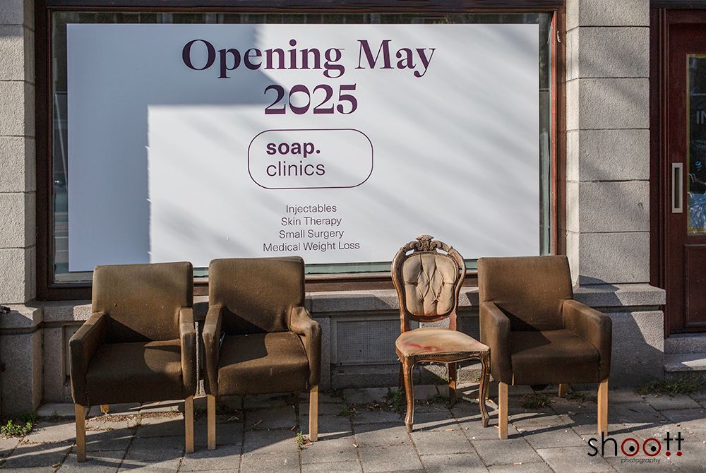 Voor een winkelraam met een bord waarop de opening van zeepklinieken in mei 2025 wordt aangekondigd staan vier stoelen op de stoep.