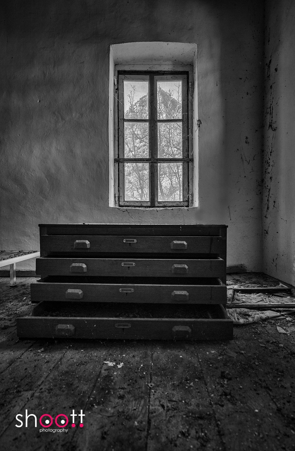 Een zwart-witfoto van een oude commode met open lades, die onder een raam staat in een verweerde, verlaten kamer.