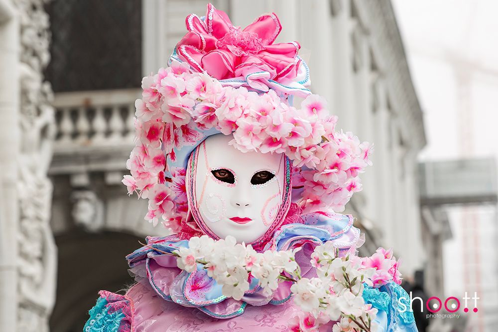 Een persoon met een Venetiaans masker en een kostuum bedekt met roze en blauwe bloemversieringen.