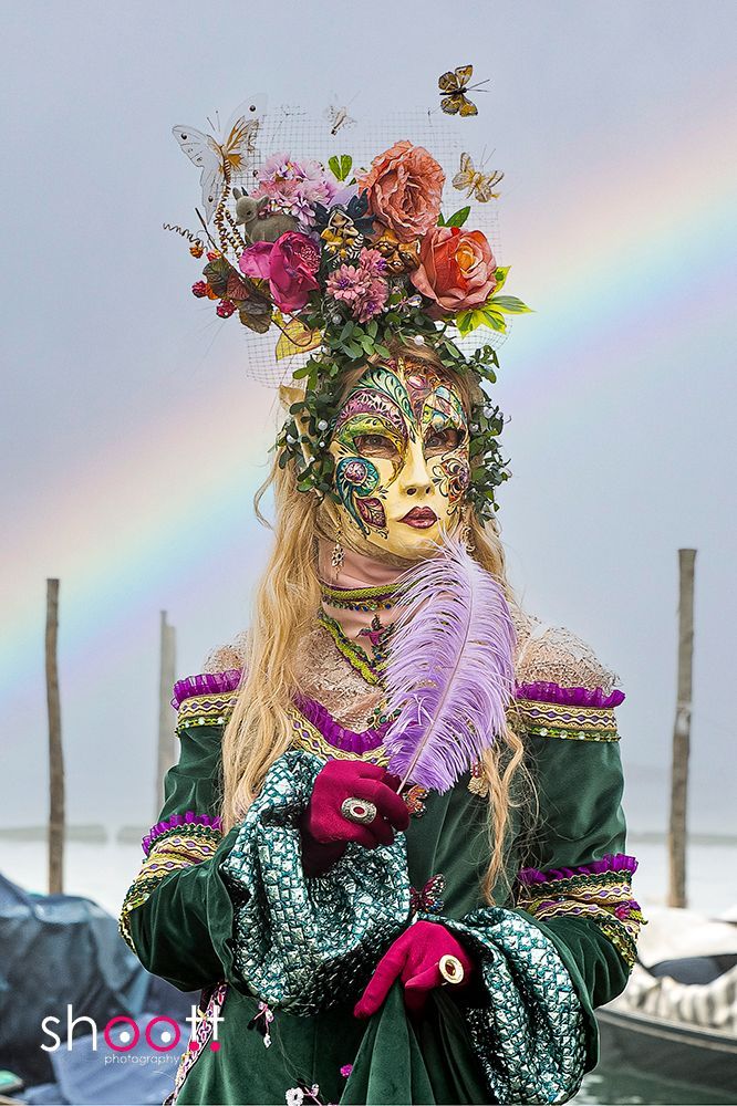 Een persoon met een sierlijk bloemenmasker en een fluwelen jurk houdt een paarse veer vast, staand in de buitenlucht met een regenboog aan de hemel.