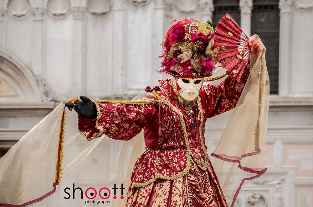 Een persoon in een rood-goud Venetiaans masker en kostuum houdt een sluier vast terwijl hij poseert voor een stenen gebouw.