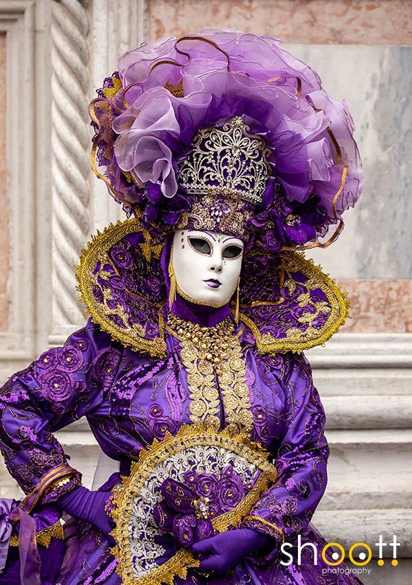 Een persoon in een weelderig, rijk versierd paars Venetiaans carnavalskostuum met masker, die een bijpassende waaier vasthoudt.