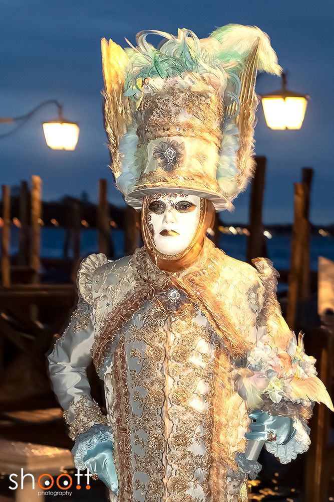 Een persoon, gekleed in een sierlijk, lichtblauw en goudkleurig Venetiaans carnavalskostuum en een versierde hoed, staat aan de waterkant van het San Marcoplein in de schemering.