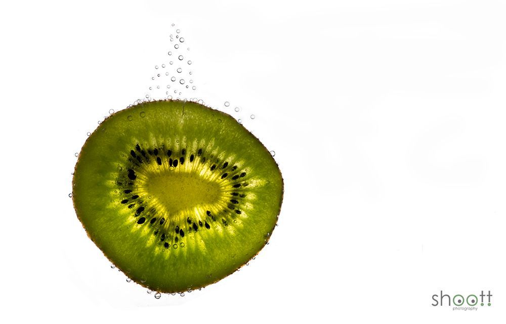 Een schijfje groene kiwi ondergedompeld in water, omgeven door kleine opstijgende luchtbelletjes tegen een witte achtergrond.