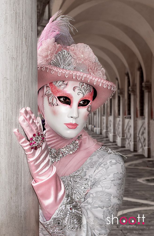 Een persoon in een decoratief roze en zilveren Venetiaans maskeradekostuum poseert tegen een pilaar in een gang.