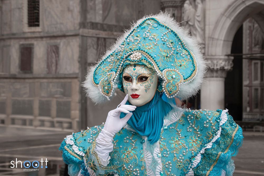 Een persoon in een decoratief blauw-goud Venetiaans carnavalskostuum met masker poseert met een hand tegen de wang.