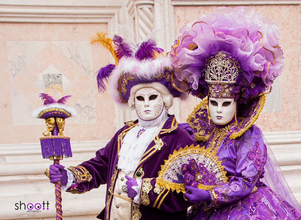 Twee mensen in uitbundige paarse, gouden en witte Venetiaanse carnavalskostuums en maskers poseren tegen een stenen muur.