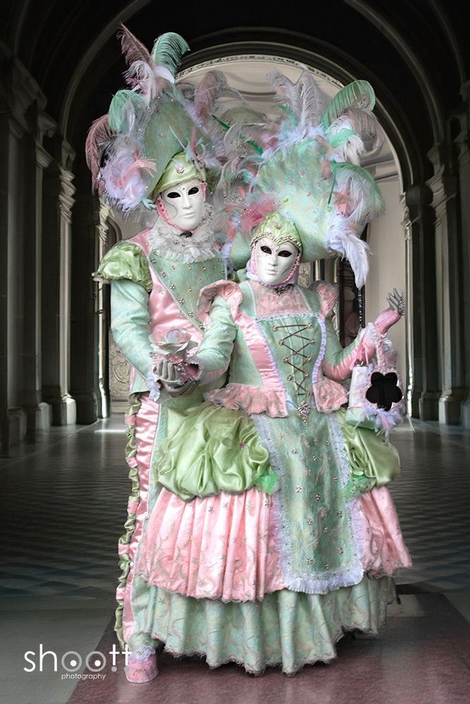 Twee figuren in sierlijke, mintgroene en lichtroze Venetiaanse carnavalskostuums en maskers staan ​​in een gang met zuilen.