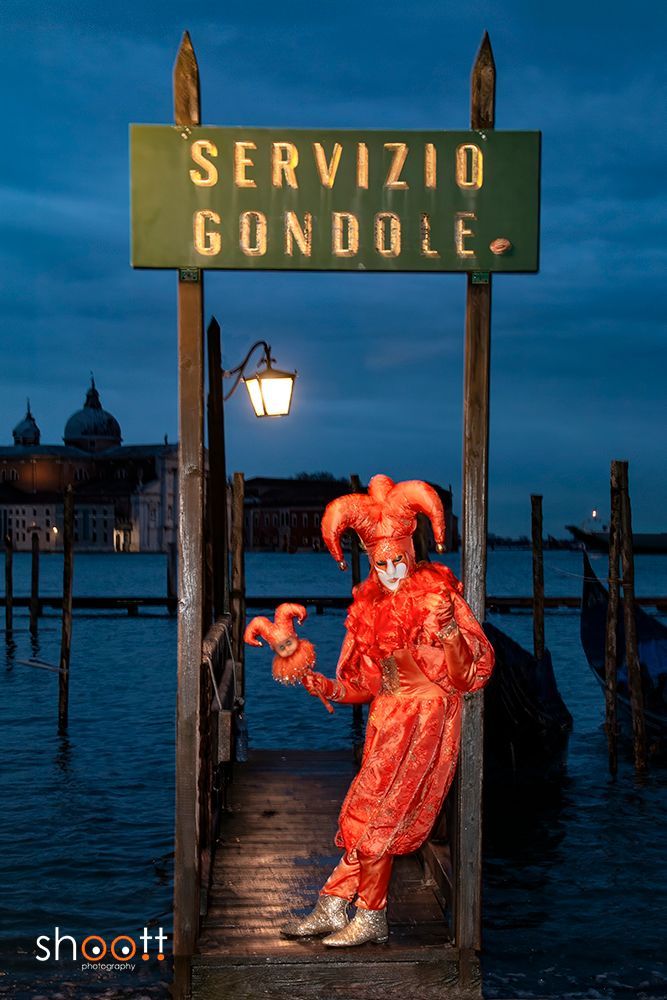 Een gemaskerde nar in een felrood kostuum staat bij schemering op een houten pier in Venetië, onder een bord met de tekst 