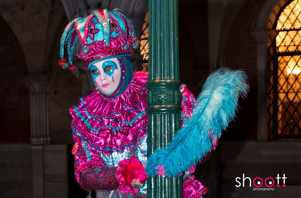 Een persoon in een felroze en blauw narrenkostuum met masker poseert achter een groene lantaarnpaal in een schemerig verlichte, gewelfde ruimte.