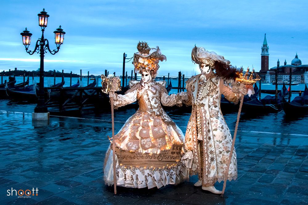 Twee mensen in sierlijke, wit-gouden carnavalskostuums van Venetië staan ​​bij schemering aan de waterkant van het San Marcoplein, vlakbij een lantaarnpaal. De jurk vertoont een driemaster zeilschip.