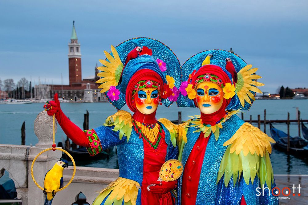 Twee mensen in uitbundige, kleurrijke Venetiaanse maskers en kostuums met papegaaienmotieven poseren aan het water in Venetië.