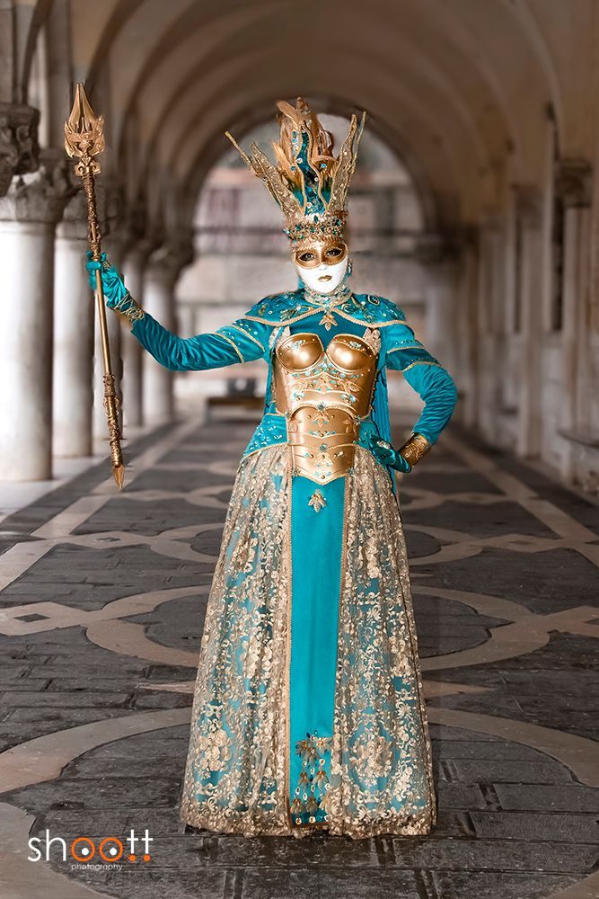 Een in een rijk versierde jurk in turquoise en goud met een uitgebreide hoofdtooi draagt ​​een staf in een gang met zuilen.
