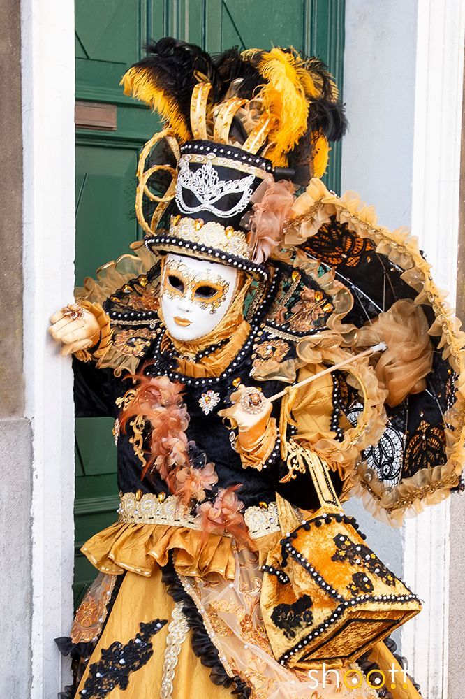 Een persoon in een zwart-goud Venetiaans maskeradekostuum met een hoed met veren en een bijpassende parasol, poseert in een deuropening.
