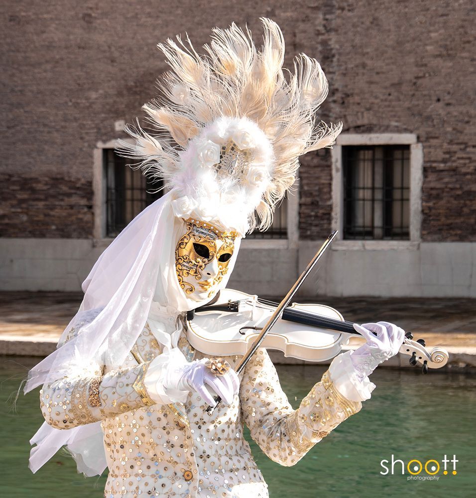 Een artiest met een gouden masker en een uitbundig wit kostuum bespeelt een witte viool nabij een kanaal bij het arsenaal in Venetië.