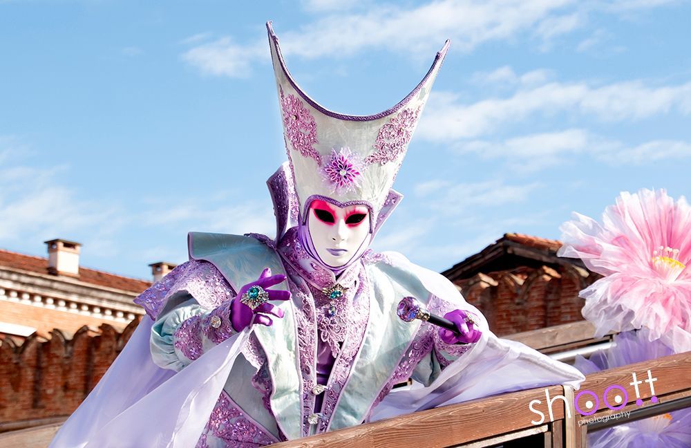 Een artiest, gekleed in een uitgebreid lavendel-wit Venetiaans masker en kostuum met een hoge, puntige hoofdtooi, staat op de brug bij het arsenaal in Venetië.