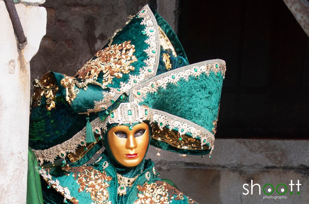 Een persoon in een sierlijk, smaragdgroen en goudkleurig Venetiaans carnavalskostuum met een bijpassende, uitgebreide hoofdtooi en masker.