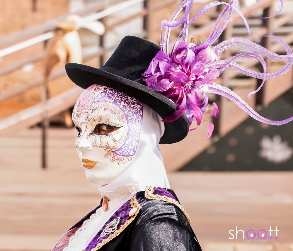 Een gemaskerde figuur met een zwarte hoge hoed versierd met paarse bloemen en veren, gekleed in een paars-zwarte outfit tijdens het carnaval van Venetië