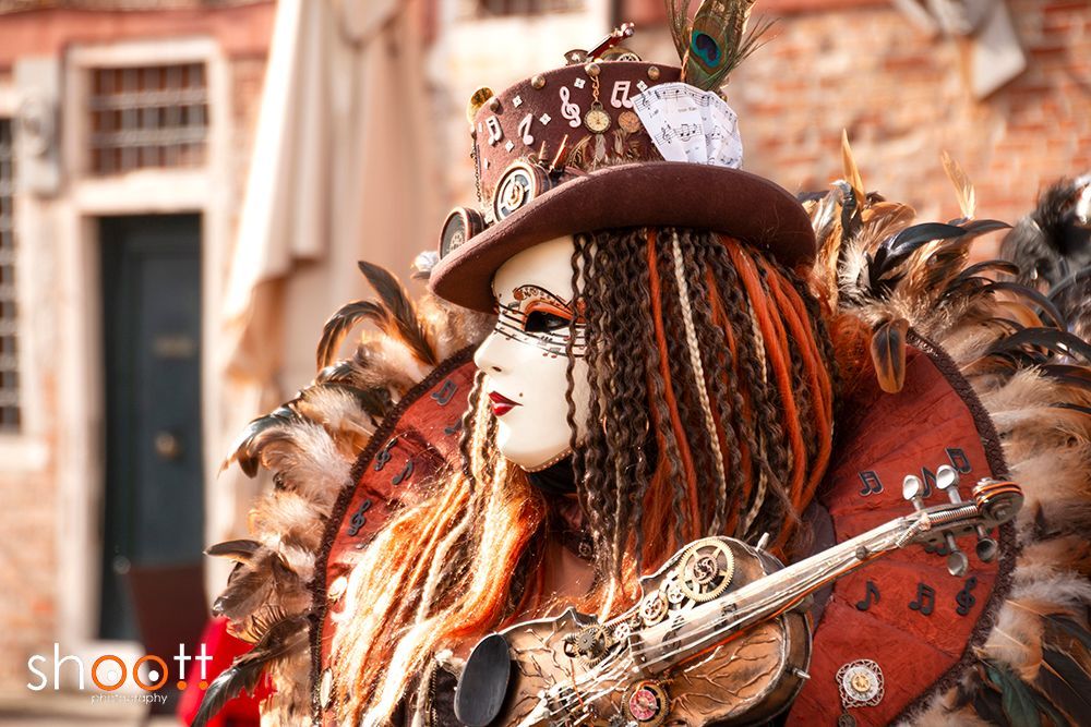 Een persoon met een theatraal Venetiaans masker, een steampunkhoed en een gevederd kostuum houdt een viool vast tegen een wazige straatachtergrond.
