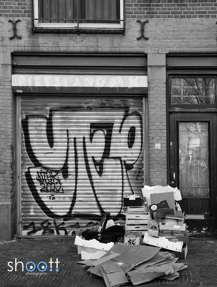 Een zwart-witfoto van een winkelpui met graffiti op een metalen rolluik en een stapel kartonnen dozen ervoor.