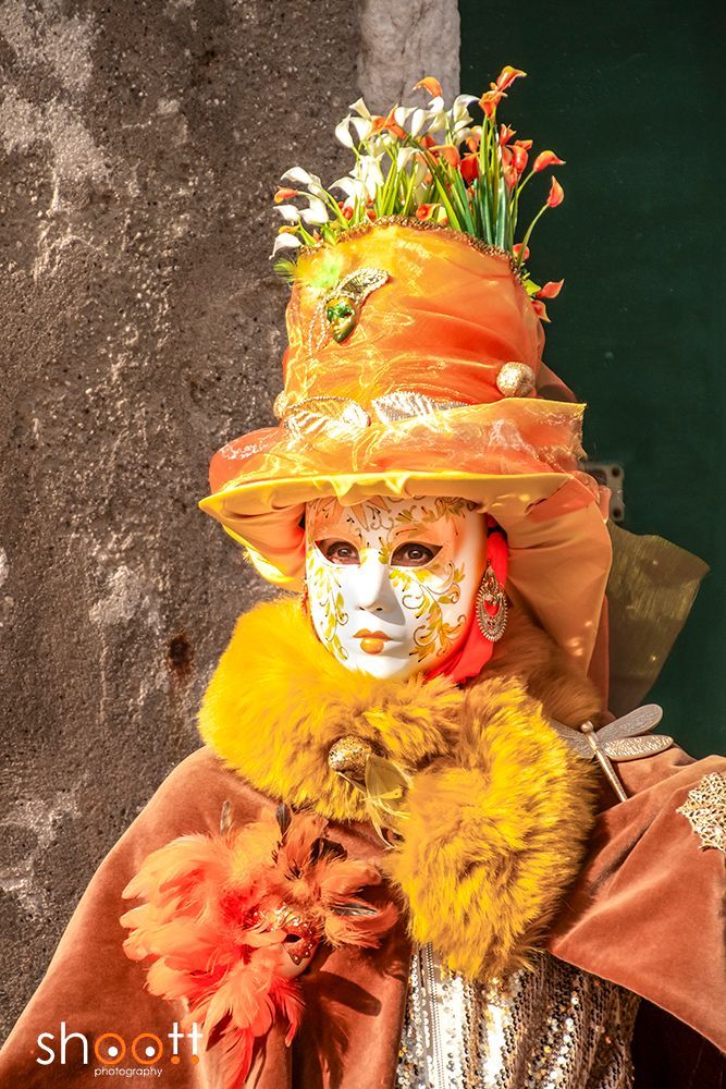 Een persoon met een decoratief gouden masker en een oranje hoge hoed versierd met bloemen en een gele bontkraag tijdens het carnaval van Venetië.