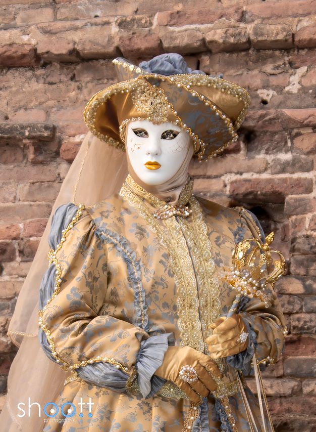 Een gemaskerde figuur in een sierlijk, goudkleurig historisch kostuum en een breedgerande hoed staat voor een rustieke bakstenen muur.