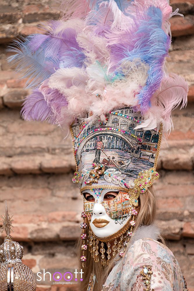 Een persoon draagt ​​een uitgebreid Venetiaans masker, een hoge, decoratieve kroon en een grote pluim van pastelkleurige veren tegen een bakstenen achtergrond.