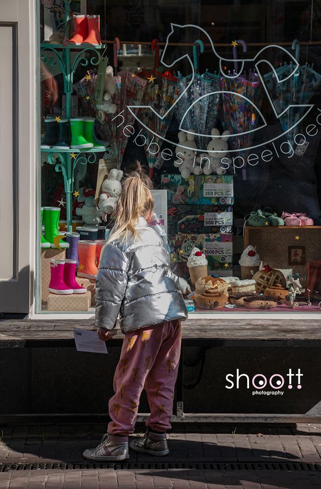 Een kind in een zilverkleurige gewatteerde jas en roze broek staat voor een speelgoedwinkel en kijkt door de etalage.