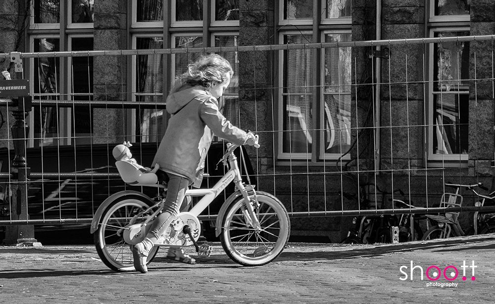Een kind fietst op een fiets met een poppenzitje achterop, langs een gebouw met grote ramen en een metalen hek.